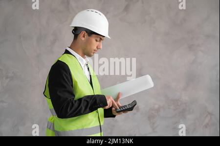 The cost of a new home. Muslim young man with drawings and a calculator isolated on grey background. Estimate of construction. A Builder in a white Stock Photo