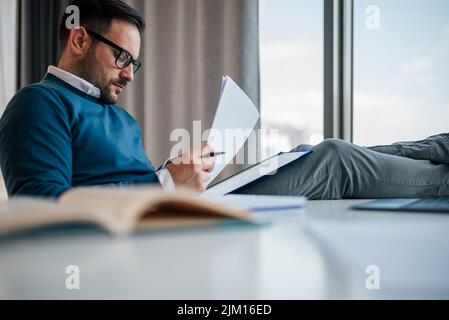 Focused professional analyzing documents. Male entrepreneur is wearing smart casuals while sitting at desk. He is working in corporate workplace. Stock Photo