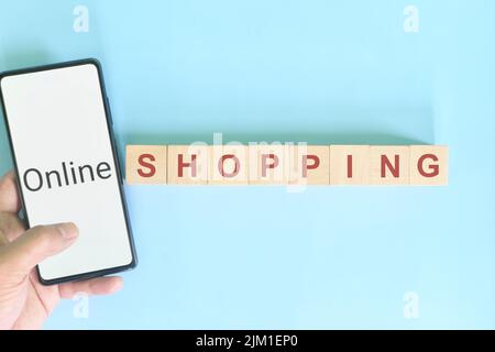 Online shopping concept. Hand holding mobile phone beside wooden blocks in blue background. Stock Photo
