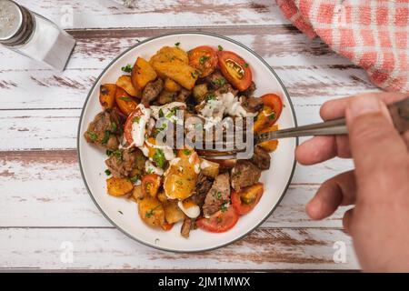 Hand taking a portion of beef chunks sauteed with cassava or manioc, chives, pepper, lemon and cherry tomato on a rustic surface. Top view Stock Photo