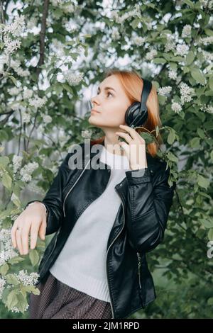 A red-haired teenage girl listens to music in headphones, configured via the phone. The time of spring flowering of cherry orchards Stock Photo