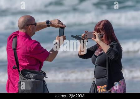 A couple of visitors using their mobile phones to photograph each other on Fistral Beach in Newquay in Cornwall in the UK. Stock Photo