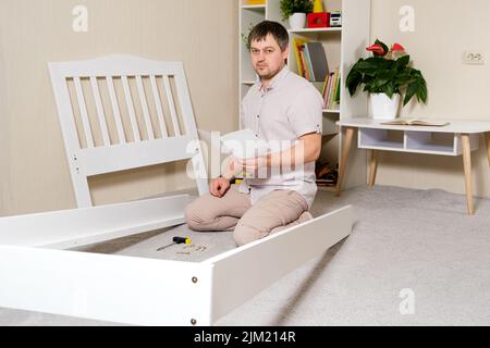 A man with instructions for assembling furniture is going to assemble a children's white wooden crib in a children's bedroom. Stock Photo