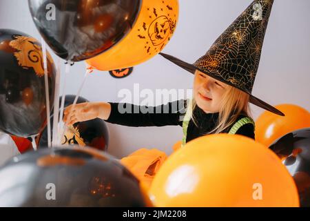 Children's Halloween - a girl in a witch hat and a carnival costume with airy orange and black balloons at home. Ready to celebrate Halloween Stock Photo