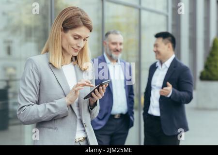 A beautiful and successful business woman is using the phone outside the office, on the photo are male colleagues talking, a successful business team is on a break Stock Photo