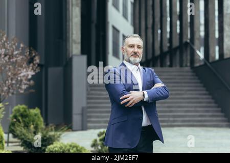 Portrait of successful thinking experienced senior businessman, gray haired male boss in business suit outside office, looking at camera with crossed arms Stock Photo