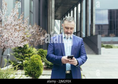 Senior experienced gray-haired businessman outside office, smiling and happy using phone, boss reading and looking at smartphone screen, walking near building Stock Photo