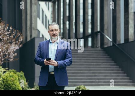 Successful senior investor with phone outside office building, businessman in business suit using smartphone and looking away dreamily Stock Photo