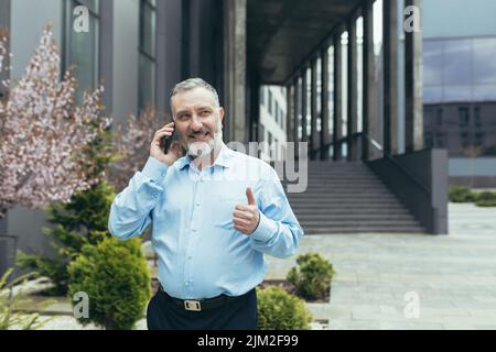 Senior gray-haired businessman in a shirt outside an office building talking on the phone, walking around the city Stock Photo