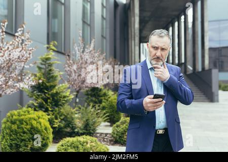 pensive senior and experienced man outside office reading news on phone, businessman outside office using smartphone while walking near building Stock Photo