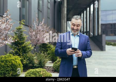Senior experienced gray-haired businessman outside office, smiling and happy using phone, boss reading and looking at smartphone screen, walking near building Stock Photo