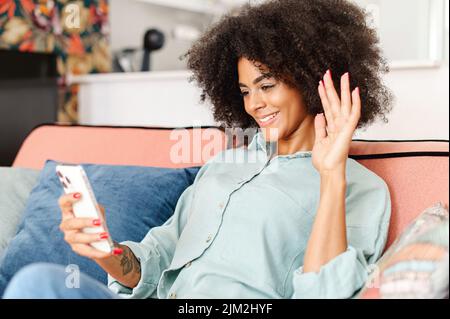 Happy African-American female using mobile app on smartphone for making video call, waving hand and greeting friends or family, talking via video connection, stay in touch on the distance Stock Photo