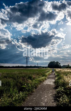 Old Woman Walks On Gravel Path Through Rural Landscape With Heavy Rainclouds And Energy Pylon Stock Photo