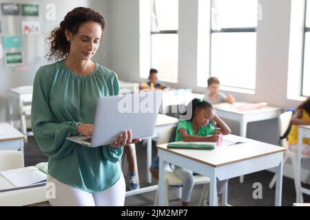 Caucasian young female teacher using laptop with multiracial elementary students in background Stock Photo
