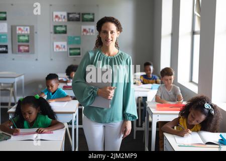 Portrait of caucasian young female teacher standing by multiracial students studying at desk Stock Photo