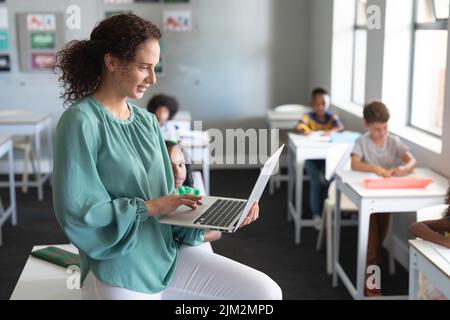 Caucasian young female teacher using laptop while sitting at desk with multiracial students studying Stock Photo