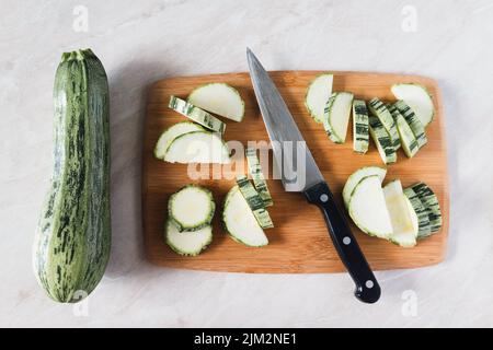 Cutting zucchini on wooden board, whole squash, knife and slices, overhead view Stock Photo