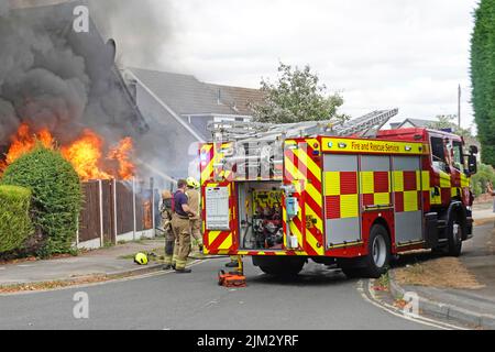 Flame & black smoke from house fire as firemen arrive in second fire brigade engine tender & crew get to work in residential street Essex England UK Stock Photo