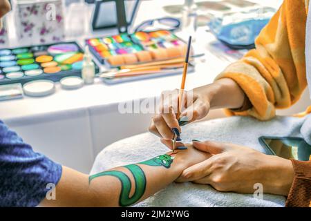 Young girl artist draws a dragon on child's hand. Painting skin with paints, close-up, without faces. Stock Photo