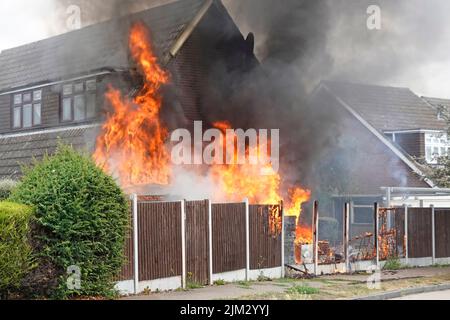 Flames from assorted household items stored outdoors beside timber boundary fence ignited in scorching summer heat setting fire against house wall UK Stock Photo