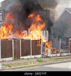 Flames from assorted household items stored outdoors beside timber boundary fence ignited in scorching summer heat setting fire against house wall UK Stock Photo