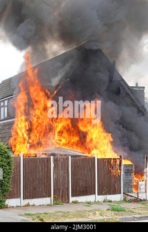 Flames from assorted household items stored outdoors beside timber boundary fence ignited in scorching summer heat setting fire against house wall UK Stock Photo