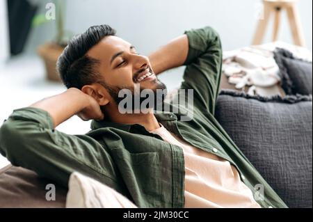 Relaxed satisfied calm mixed race guy in casual shirt, resting on the sofa in cozy living room with his eyes closed, throwing his hands behind his head, dreaming of vacation, resting from work, smiles Stock Photo