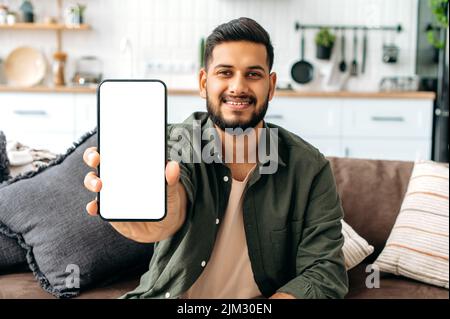 Mock-up and copy-space concept. Positive indian or arabian guy in casual wear, sits on sofa in living room, shows smart phone with empty white mock-up screen, for advertisement or presentation, smiles Stock Photo