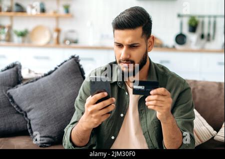 Focused Indian or Arabian attractive guy, sit on the sofa in the living room, using a bank card and a smartphone, making online purchases, making payments, ordering delivery. Online shopping, payment Stock Photo