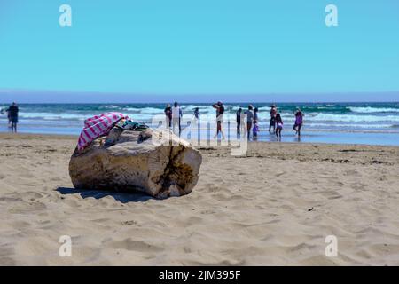 A log on the beach with a towel & flip flops on top. People swimming & walking on the beach & the ocean in the background. Pismo Beach California. Stock Photo