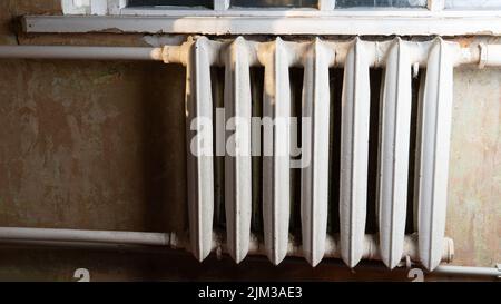 Old vintage cast iron radiator and pipes covered with white paint. Soviet retro interior. Old shabby walls without wallpaper behind the radiator. Heating equipment at home.  Stock Photo