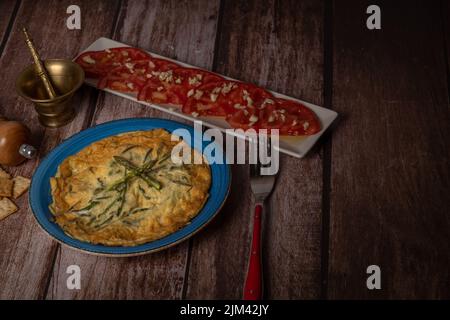 wild asparagus omelette on a blue plate and tomato salad with garlic on a wooden mortar and pestle and pepper table Stock Photo