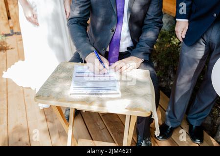A closeup shot of a groom signing a wedding contract papers on a wooden table on a sunny day Stock Photo