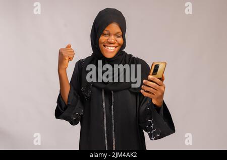 An African happy Muslim smiling excited lady using her phone and isolated on gray background Stock Photo