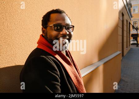 Black bearded man in sunglasses smiling while standing by wall outdoors Stock Photo