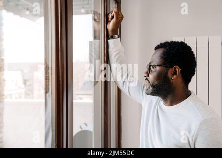Black bearded man wearing eyeglasses looking in window at home Stock Photo