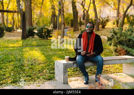 Black bearded man in sunglasses using cellphone while sitting on bench at park Stock Photo