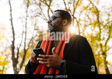 Black bearded man in sunglasses using cellphone while strolling at park Stock Photo