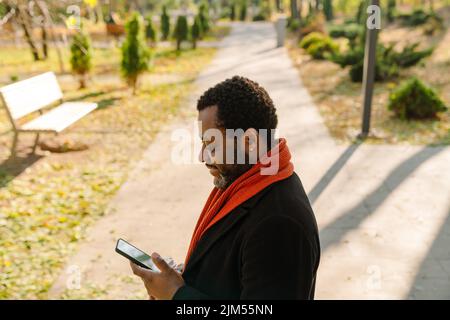 Black bearded man using cellphone and drinking coffee while strolling in park Stock Photo