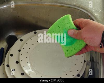 A man holding a green foam sponge for washing dirty dishes in the sink Stock Photo