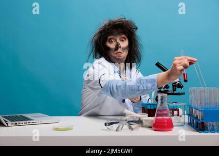 Dumb looking crazy chemist with wacky hairstyle and dirty face sitting at desk in laboratory handling glass test tubes. Mad goofy scientist holding glass beaker while looking silly at camera. Stock Photo