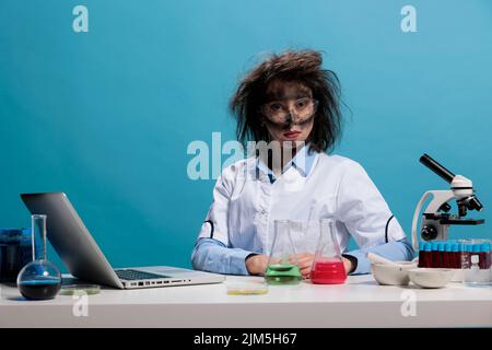 Foolish lab worker with wacky hair and dirty face sitting at desk on blue background while looking at camera. Silly looking crazy chemist with messy hairstyle after failed experiment. Stock Photo