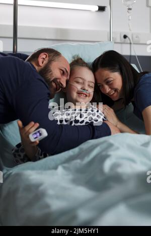 Loving parents hugging sick daughter resting in healthcare clinic pediatrics ward. Happy ill little girl hugged by caring mother and father while sitting with her on patient bed. Stock Photo