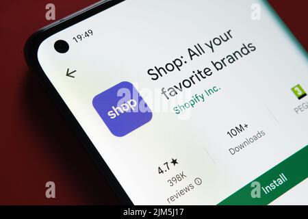 Shop app from Shopify seen in Google Play Store on the smartphone screen placed on red background. Close up photo with selective focus. Stafford, Unit Stock Photo