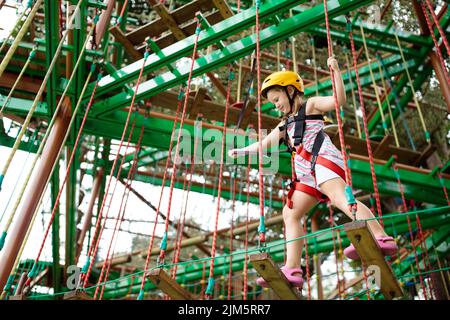 Child in forest adventure park. Kids climb on high rope trail. Agility and climbing outdoor amusement center for children. Stock Photo