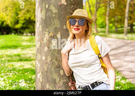 Blonde Caucasian smiling woman in sunglasses and casual clothes leaning on a tree in the park Stock Photo