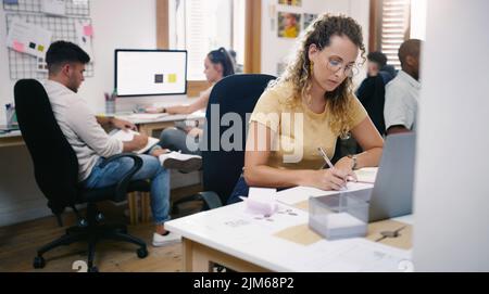 Heads down, productivity up. a young businesswoman using a laptop and making notes in a modern office. Stock Photo