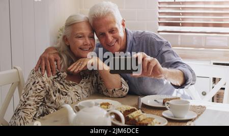 Lets capture this moment. a happy senior couple sitting together and using a cellphone to take selfies at home. Stock Photo