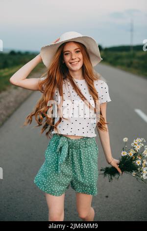 Lovely redheaded girl in sun hat, holding beautiful flowers smiling at camera while walking on the country road. Elegant model pose at nature. Stock Photo