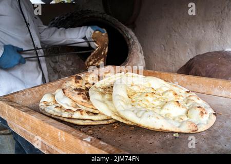 aged man makes traditional bread in old round stone oven in rural village. Large flat freshly baked tortillas lie on the table. Hot bread. Central Asi Stock Photo
